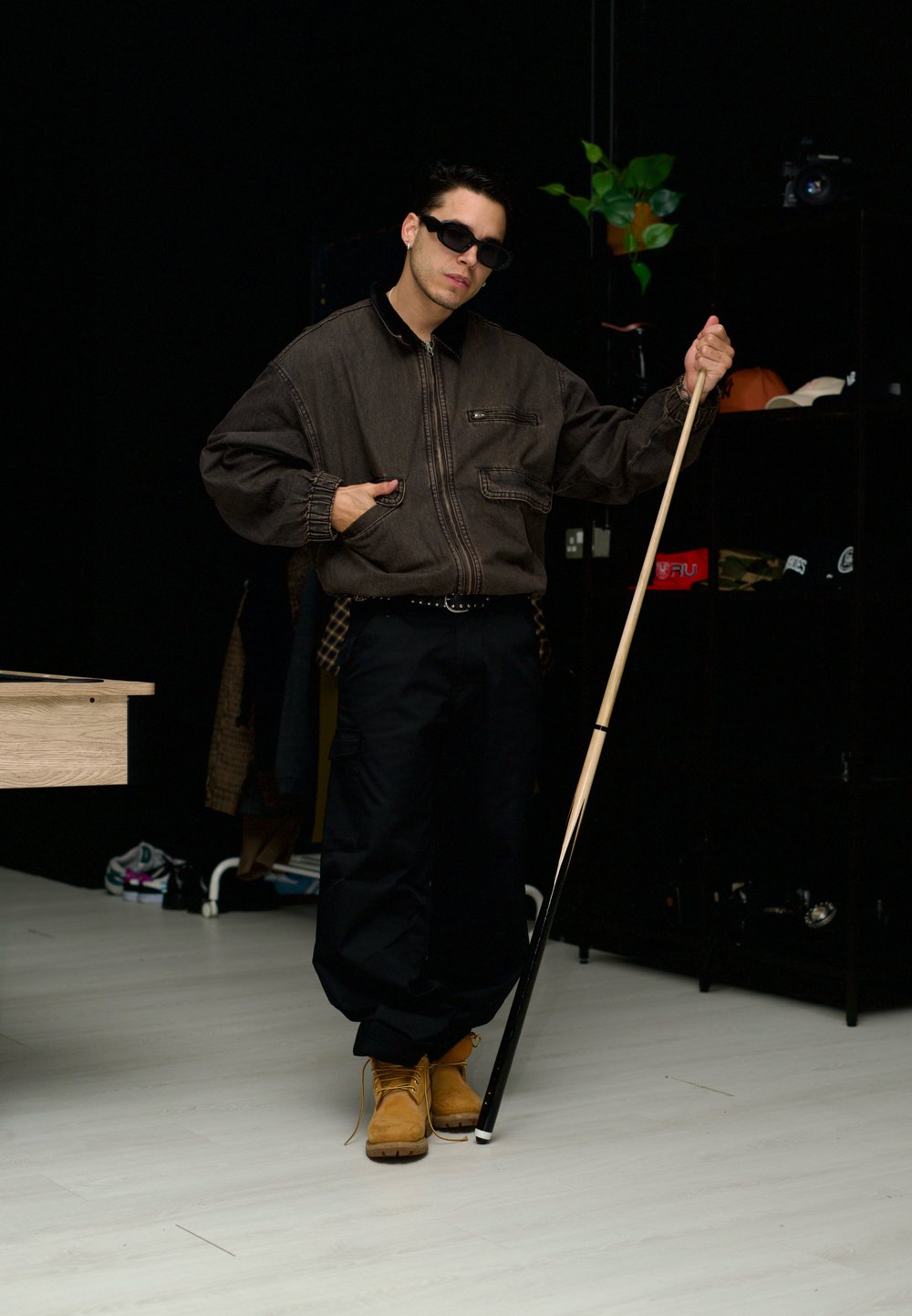 Schwarze übergroße Jacke mit Reißverschluss, schwarze Cargo-Hosen, gelbe Stiefel und Sonnenbrille. Hält einen weiß getippten Stock. Dunkler Hintergrund mit Regalen.