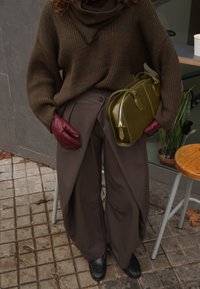 Pull marron en tricot, pantalon marron ample avec plis, gants en cuir bordeaux, sac à main vert et mocassins noirs sur une surface carrelée.