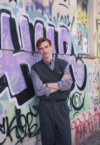 Hall halli vest maroonkaunistustega helehallil särgil. Tumedad kõrge vöökohaga püksid, seisab värvika grafitiseina taustal.