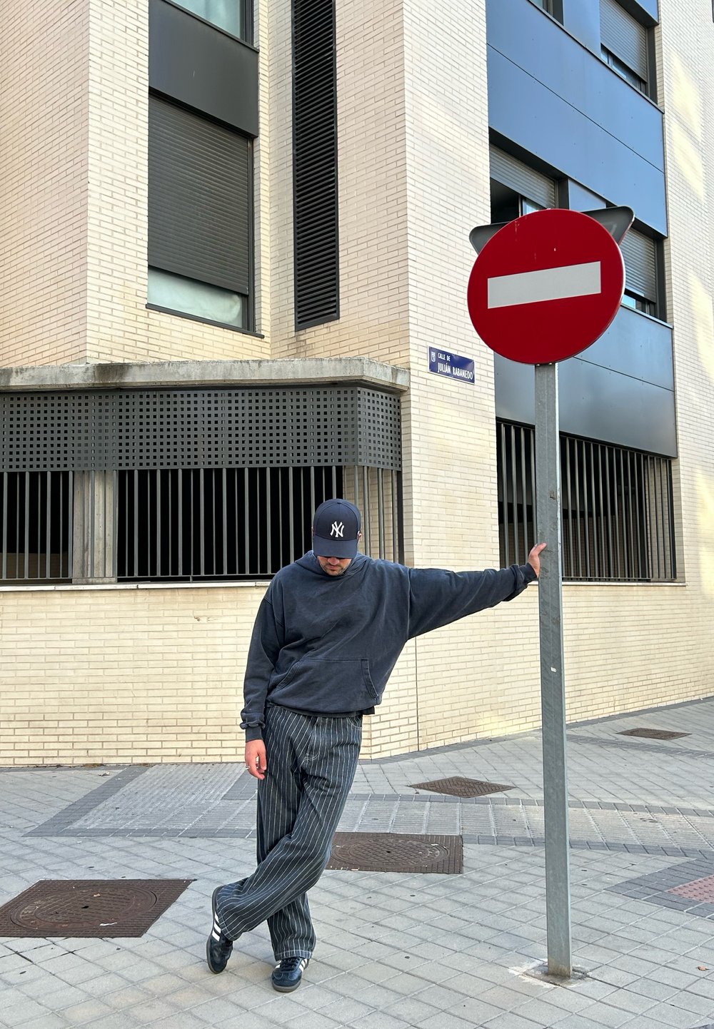 Σκούρο γκρι φαρδύ φούτερ, ριγέ παντελόνι μαύρο και γκρι, στέκεται δίπλα σε σήμα απαγόρευσης εισόδου κόκκινου χρώματος μπροστά σε ένα μπεζ τούβλινο κτίριο.