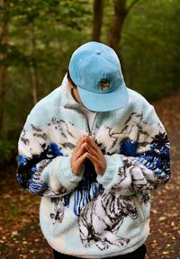 Fleece-pulloveři s tygřím vzorem v bílé, černé a modré barvě na světle modrém pozadí. Vybaven vysokým límcem a zipovým zapínáním.