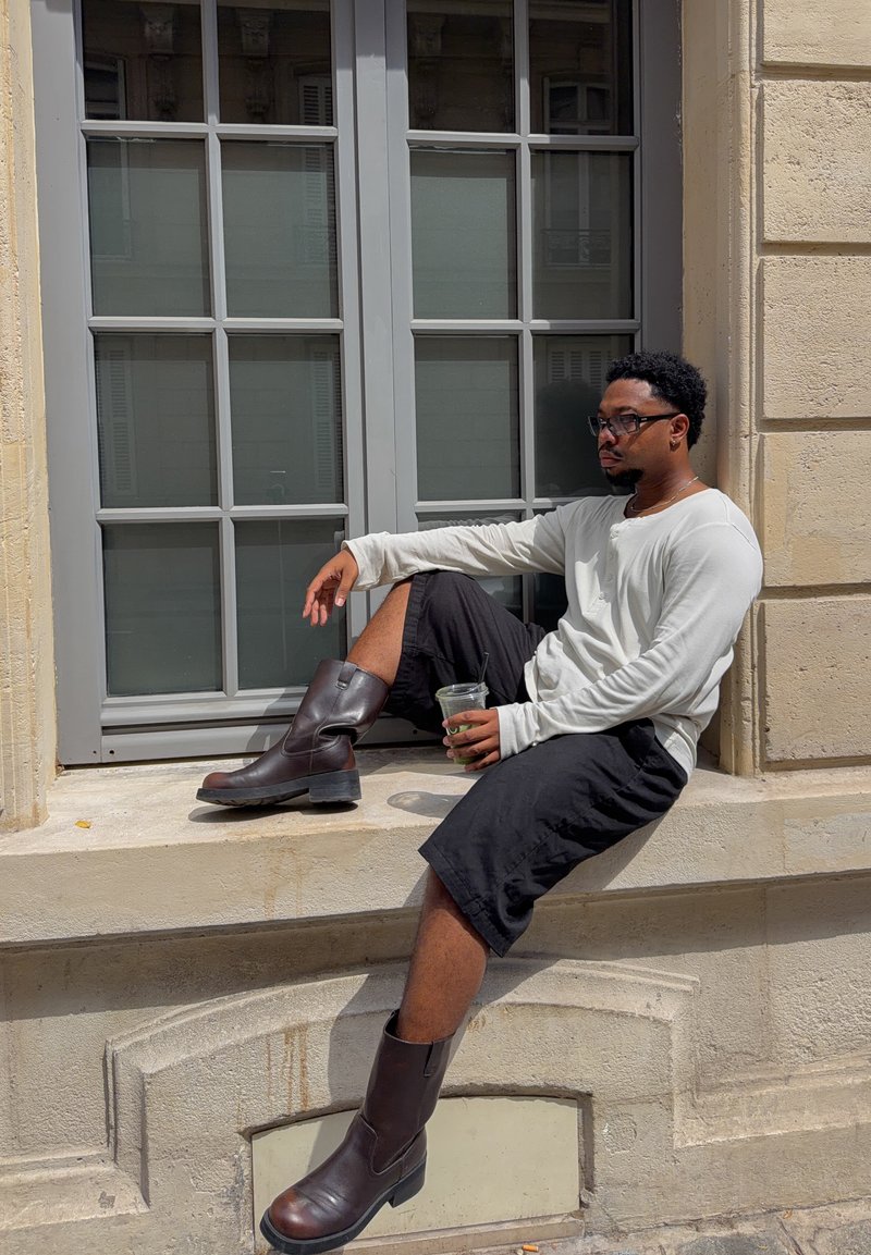 Homme portant des lunettes, une chemise blanche à manches longues, un short noir et des bottes marron, assis sur le rebord de la fenêtre tenant une boisson dans un gobelet transparent.