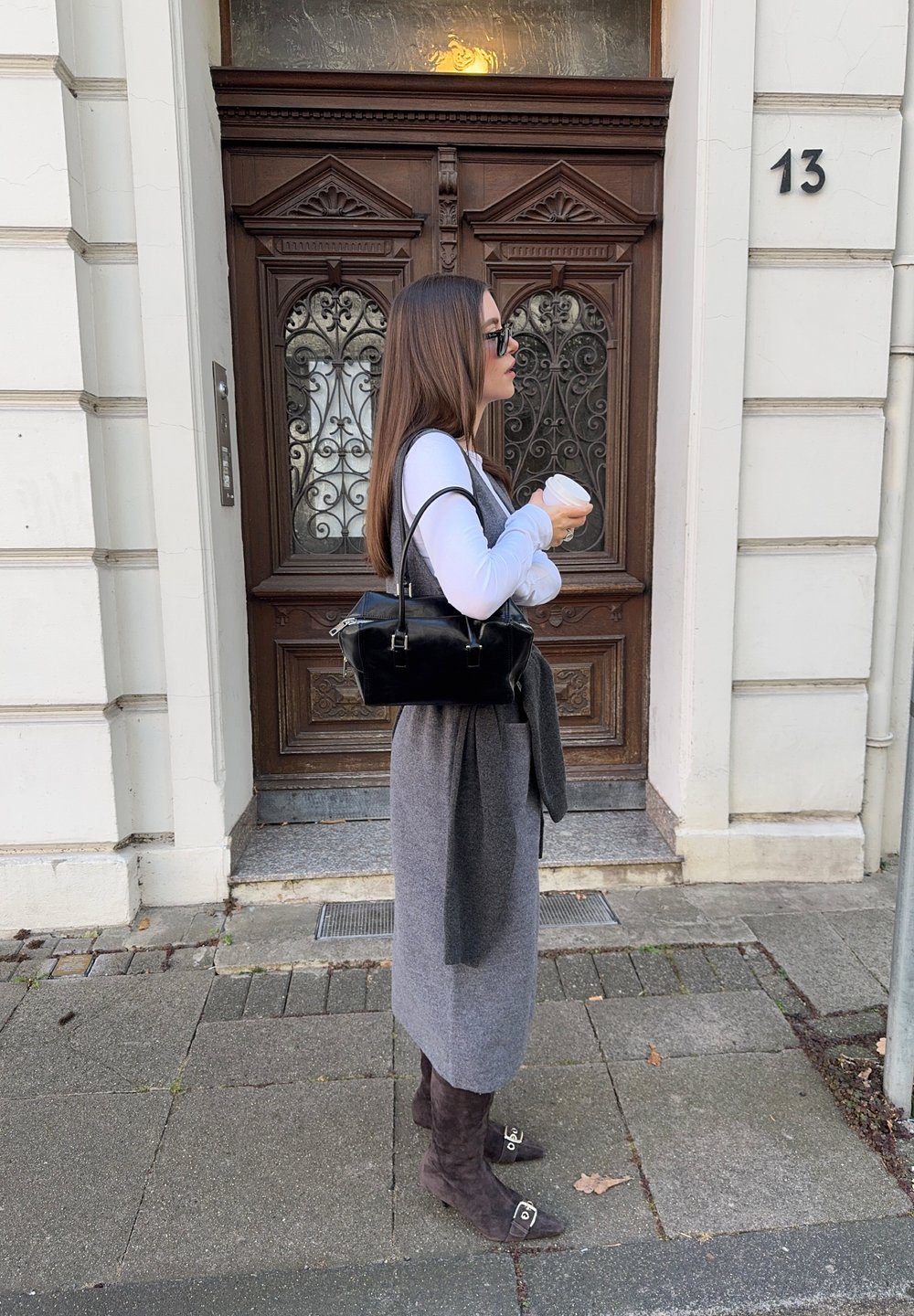 Geantă neagră, fustă gri, bluză albă cu mâneci lungi și cizme maro până la genunchi. Stă în fața unei uși din lemn ornamentate cu numărul 13.