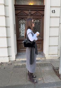 Geantă neagră, fustă gri, bluză albă cu mâneci lungi și cizme maro până la genunchi. Stă în fața unei uși din lemn ornamentate cu numărul 13.