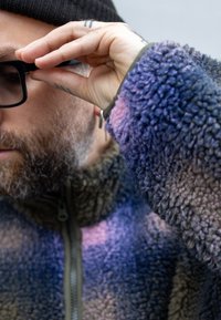 Fleecová bunda v viacfarebných textúrach purpurovej a sivej farby so zipsom. Osoba si upravuje čierne okuliare, pričom sa odhaľujú tetovania na ruke.