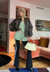 Jeune personne portant un chapeau en fourrure, une veste bomber foncée, un short rayé vert par-dessus un pantalon noir évasé, tenant un sac à main crème dans un salon.