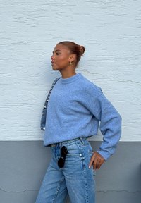 Femme aux cheveux en chignon portant un pull bleu et un jean, debout de profil contre un mur texturé blanc et gris, portant un sac à épaule clouté.