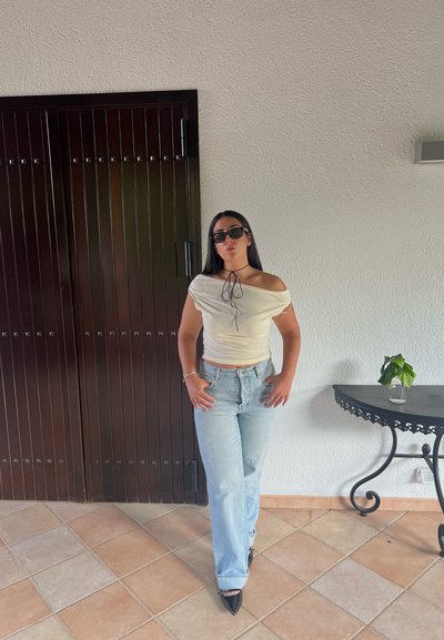 Top crema con hombros al aire, jeans azul claro, tacones negros puntiagudos, gafas de sol negras, con una planta en una mesa decorativa al fondo.