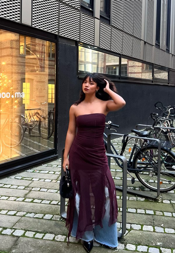 Borstloze bordeauxrode jurk met een doorschijnende overlay, gelaagd over lichtblauwe denim. Zwarte handtas en puntige zwarte hakken maken de outfit compleet.