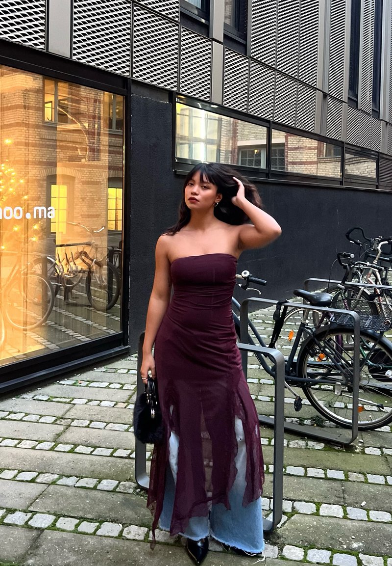 Robe bordeaux sans bretelles avec un voile transparent, superposée sur un denim bleu clair. Sac à main noir et talons noirs pointus complètent la tenue.