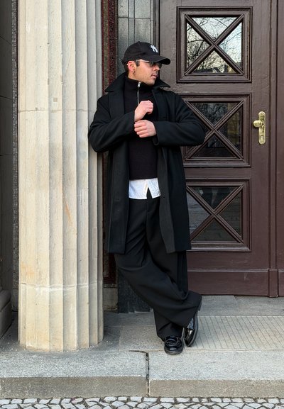 Hombre con abrigo negro, pantalones y gorra, apoyado contra una columna de piedra junto a una puerta de madera ornamentada, ajustándose la manga y mirando de lado.