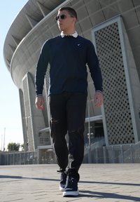 Marineblå langærmet skjorte med hvid krave, sorte slidte jeans og marineblå sneakers, parret med runde sorte solbriller. Urban indstilling.