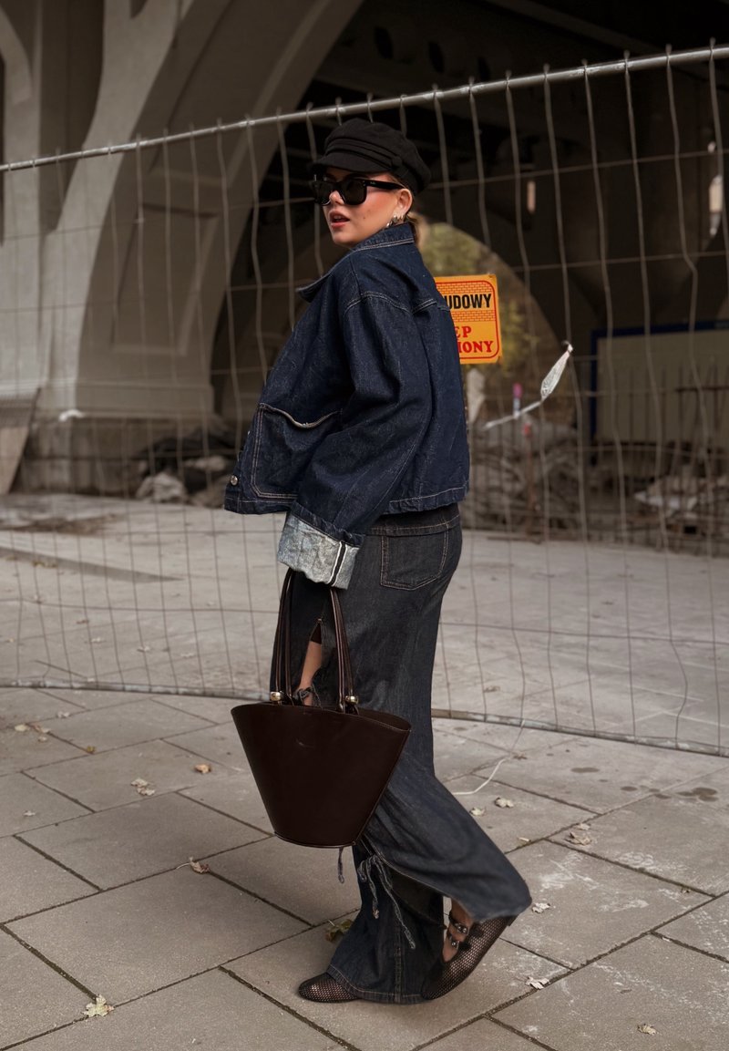 Jaqueta de ganga escura, suéter cinza, calças escuras de cintura alta com fendas laterais e uma bolsa tipo balde castanha, combinadas com sapatos de rede pretos.