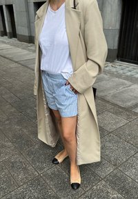 Casaco longo bege sobre uma t-shirt branca, combinado com calções azul-claro às riscas e sapatos baixos bege com pontas pretas. Bolso visível nos calções.