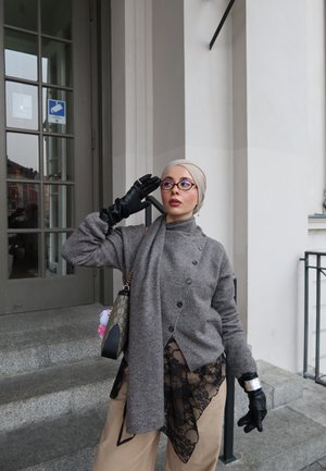 Mujer con pañuelo beige en la cabeza, gafas, suéter gris, guantes negros y pantalones marrón claro con encaje negro, posando cerca de la entrada de un edificio con escaleras.