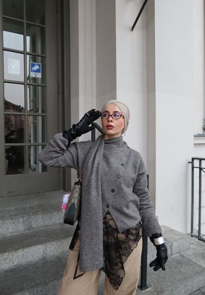 Mujer con pañuelo beige en la cabeza, gafas, suéter gris, guantes negros y pantalones marrón claro con encaje negro, posando cerca de la entrada de un edificio con escaleras.