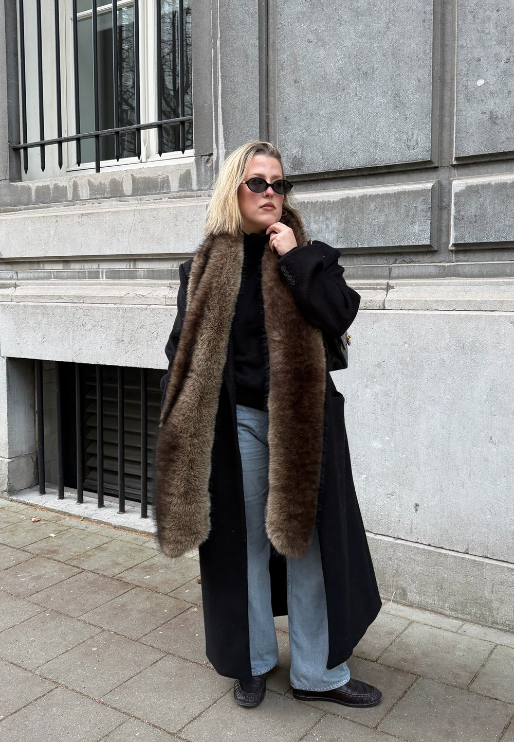 Femme portant un manteau noir avec une longue écharpe en fourrure brune, des lunettes de soleil noires, un jean bleu clair et des chaussures noires, debout sur un trottoir en ville.