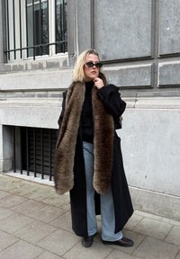 Femme portant un manteau noir avec une longue écharpe en fourrure brune, des lunettes de soleil noires, un jean bleu clair et des chaussures noires, debout sur un trottoir en ville.