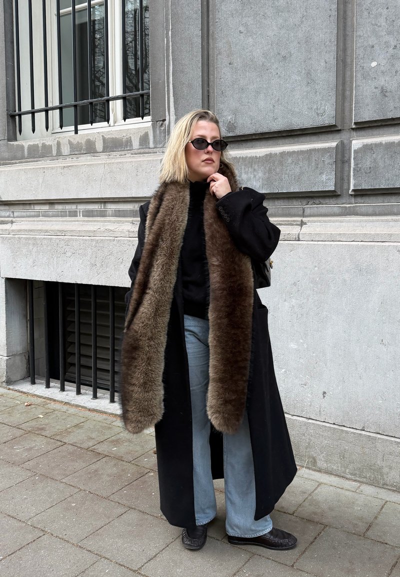 Femme portant un manteau noir avec une longue écharpe en fourrure brune, des lunettes de soleil noires, un jean bleu clair et des chaussures noires, debout sur un trottoir en ville.