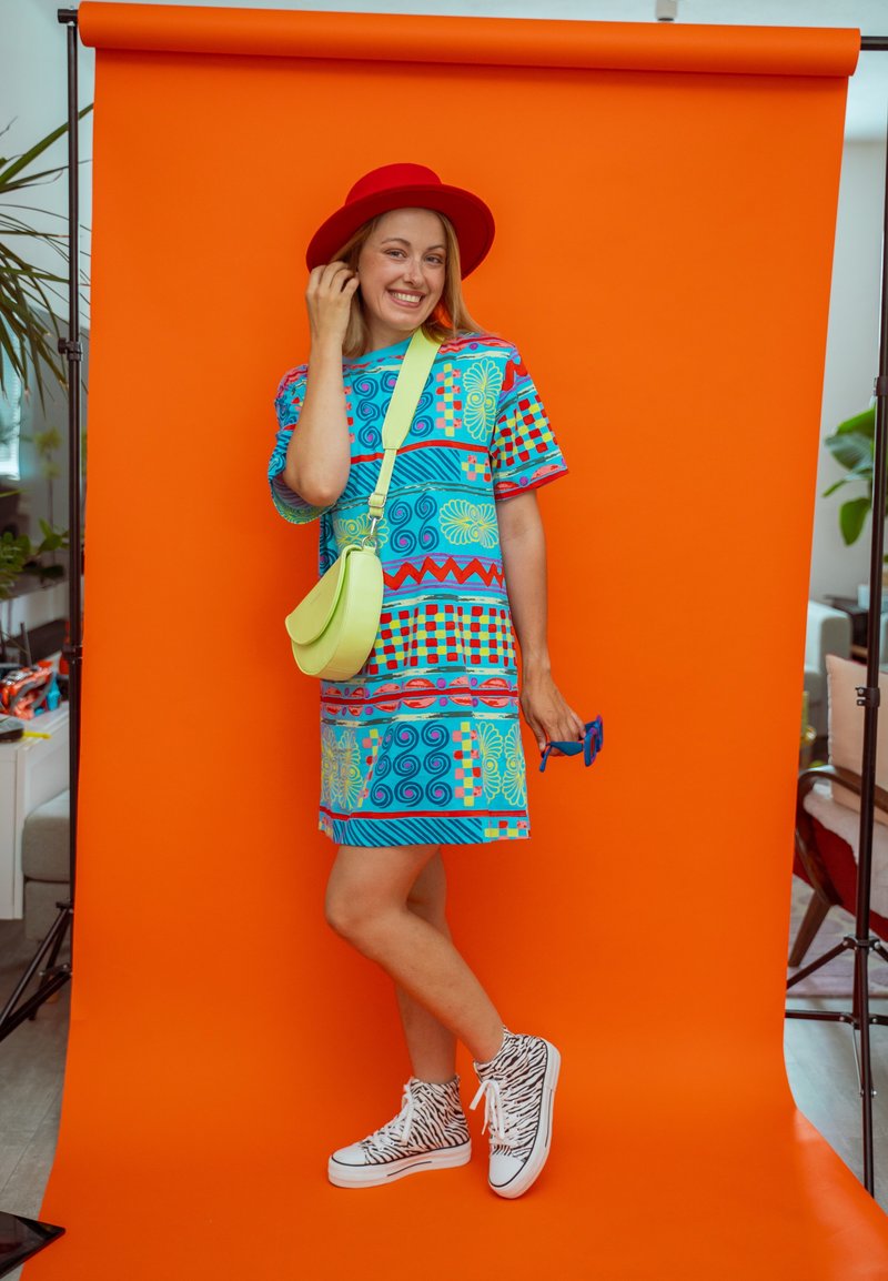 Femme souriante posant devant un fond orange, portant un chapeau rouge, une robe à motifs colorés, des baskets à imprimé zèbre, tenant des lunettes de soleil bleues et un sac vert lime.