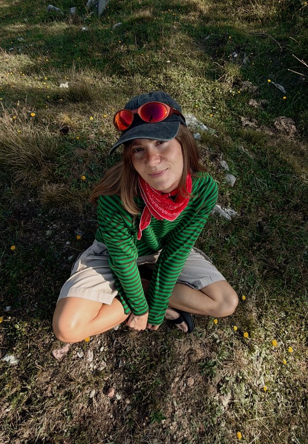 Jeune personne portant un pull vert rayé, un bandana rouge, une casquette noire avec des lunettes de soleil rouges, assise jambes croisées sur l'herbe avec de petites fleurs jaunes.