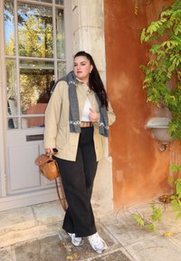 Beżowa kurtka na białej bluzce, czarne spodnie, szary szalik, brązowa torebka. W skład kompletu wchodzą sneakersy, stojąc pod pomarańczową ścianą w otoczeniu zieleni.