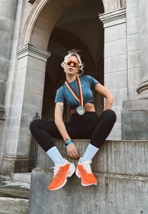 Atleta con un top corto color verde azulado y leggings negros sentado en escalones de piedra. Lleva zapatillas Brooks naranjas y gafas de sol reflectantes, con una medalla alrededor del cuello.