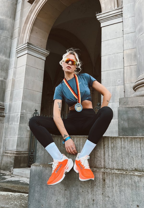 An athlete dressed in a teal crop top and black leggings sits on stone steps. They are wearing orange Brooks trainers and reflective sunglasses, with a medal around their neck.