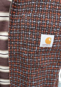 Brunt og orange ternet stof med blå accenter, indeholdende et gråt stribet element og et Carhartt-logo på en glat overflade.