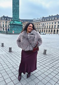 Casaco de pelúcia falsa em cinza claro, saia justa bordeaux, botas de tornozelo brilhantes na cor bordô e mala; praça da cidade com um fundo de coluna histórica.