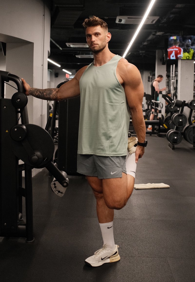 Homme musclé en tenue de sport étirant le quadriceps en tenant un pied derrière lui, debout à côté d’une machine d’exercice.