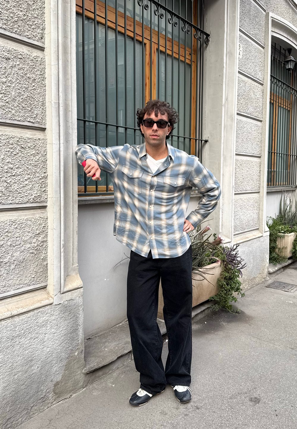 Light blue and white plaid button-up shirt, black trousers, and black sneakers. Standing in front of a building with decorative windows.