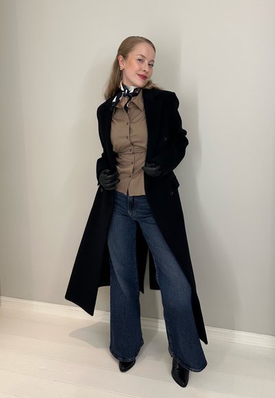 Mujer con abrigo largo negro, camisa beige abotonada, vaqueros anchos azules, guantes negros, botas negras puntiagudas y pañuelo de seda alrededor del cuello.