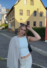 Biała dopasowana bluzka, beżowa lniana kurtka zawieszona na ramionach i jasnobeżowa spódnica. Czarna torba, trzymająca okulary przeciwsłoneczne, stojąca na wybrukowanej ulicy w pobliżu budynków.