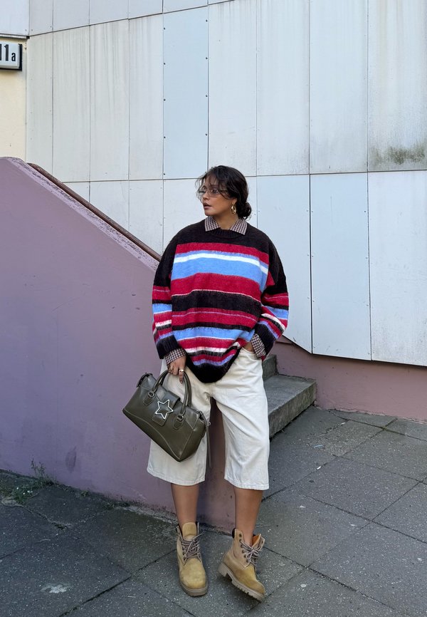 Une femme se tient près d’un escalier. Elle porte un pull rayé, un short blanc et des bottes, et tient un sac à main vert foncé.