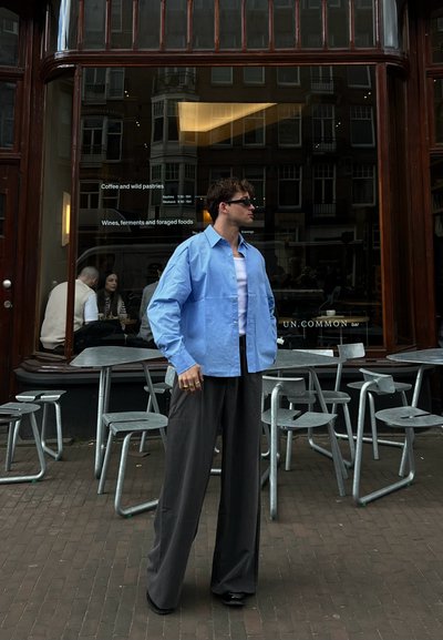 Camisa oversized azul clara, top negro de tirantes y pantalones anchos negros. Gafas de sol, anillos, superficies texturizadas y ambiente de café al aire libre.