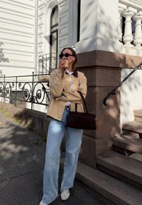 Giacca beige chiaro con colletto marrone, camicia bianca, jeans azzurri e sneakers bianche. Tiene in mano una tazza di caffè marrone, in piedi accanto a un muro di pietra.