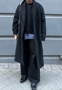 Černý vlněný kabát, texturovaný černý svetr, modrá kostkovaná košile a volné černé kalhoty. Celkový vzhled doplňují černé kožené boty.
