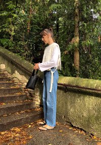 Top branco texturizado sobre uma camisa de manga longa, jeans de ganga azul de cintura alta, bolsa tote preta e chinelos numa escadaria de pedra florestal.