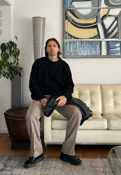 Suéter negro de punto, pantalones marrones a cuadros y zapatos negros. Sentado en un sofá de cuero blanco con un jarrón alto y texturizado y una obra de arte detrás.