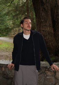 Homme portant une veste zippée bleu marine foncé sur un pull en maille, pantalon gris clair, debout contre un mur en pierre, entouré d'arbres.