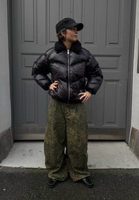 Pufasta crna jakna s mekanim ovratnikom i srebrnim patentnim zatvaračem, uparena s prevelikim zelenim hlačama s uzorkom i crnim cipelama, ispred sive vrata.