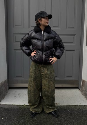 Chaqueta negra acolchada con cuello de pelo y cremallera plateada, combinada con pantalones verdes con estampado grande y zapatos negros, frente a una puerta gris.