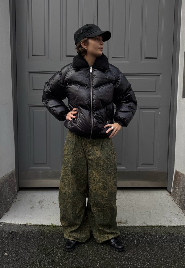 Giacca nera imbottita con colletto peloso e cerniera argentata, abbinata a pantaloni verde oversize con motivi e scarpe nere, davanti a una porta grigia.