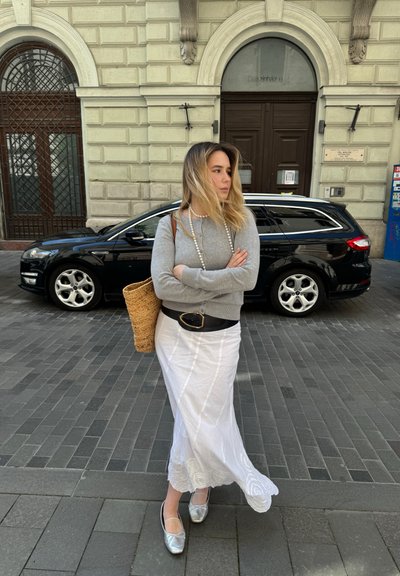 Una mujer con un suéter gris lleva una falda blanca y fluida con bordado floral, zapatos planos plateados y un bolso de mimbre, con un fondo de ciudad.