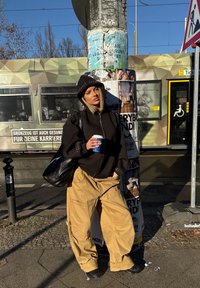 Schwarze Zip-Hoodie mit Nietenakzenten, beige baggy Cargo-Hosen, hält eine blaue Kaffeetasse, lehnt sich gegen einen Laternenpfahl mit Plakaten.