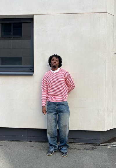 Camisa de rayas rojas y blancas de manga larga, jeans azules holgados, de pie contra una pared de color claro con un marco de ventana gris.