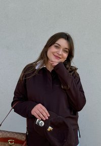 Jonge vrouw met bruin haar glimlacht vriendelijk, draagt een donker shirt met lange mouwen, houdt een bruine pet vast en draagt een bruine handtas met een patroon.