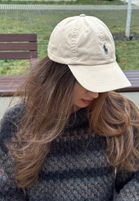 Γυναίκα με μακριά καστανά μαλλιά, που φοράει μπεζ μπέιζμπολ καπέλο Polo και σκούρο πλεκτό πουλόβερ, καθισμένη σε παγκάκι σε εξωτερικό χώρο.