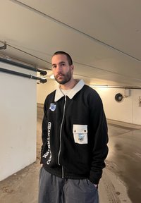 Μαύρο φούτερ με φερμουάρ και άσπρο γιακά και τσέπη, διακοσμημένο με μπαλώματα και κείμενο. Γκρίζο παντελόνι, στέκεται σε υπόγειο χώρο στάθμευσης.